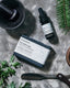 A Lavender & Black bar of soap and small dropper bottle laying on a marble stone surface, surrounded by  botanicals and a shaving tool