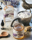 Open jar of lavender honey, bottle of lavender syrup, and small dropper bottle, on a stone marble table surrounded by wooden tools and fresh lavender sprigs