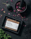 A brown soap bar with white label on a charcoal black surface, surrounded by pink dried flower petals