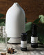 An essential oil and spray bottle on a stone surface, place in front of a white diffuser and fresh leaves