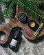 small shave oil bottle and an open round wild resin hand salve container, on an abstract wooden cutting board, surrounded by fresh douglas fir branches